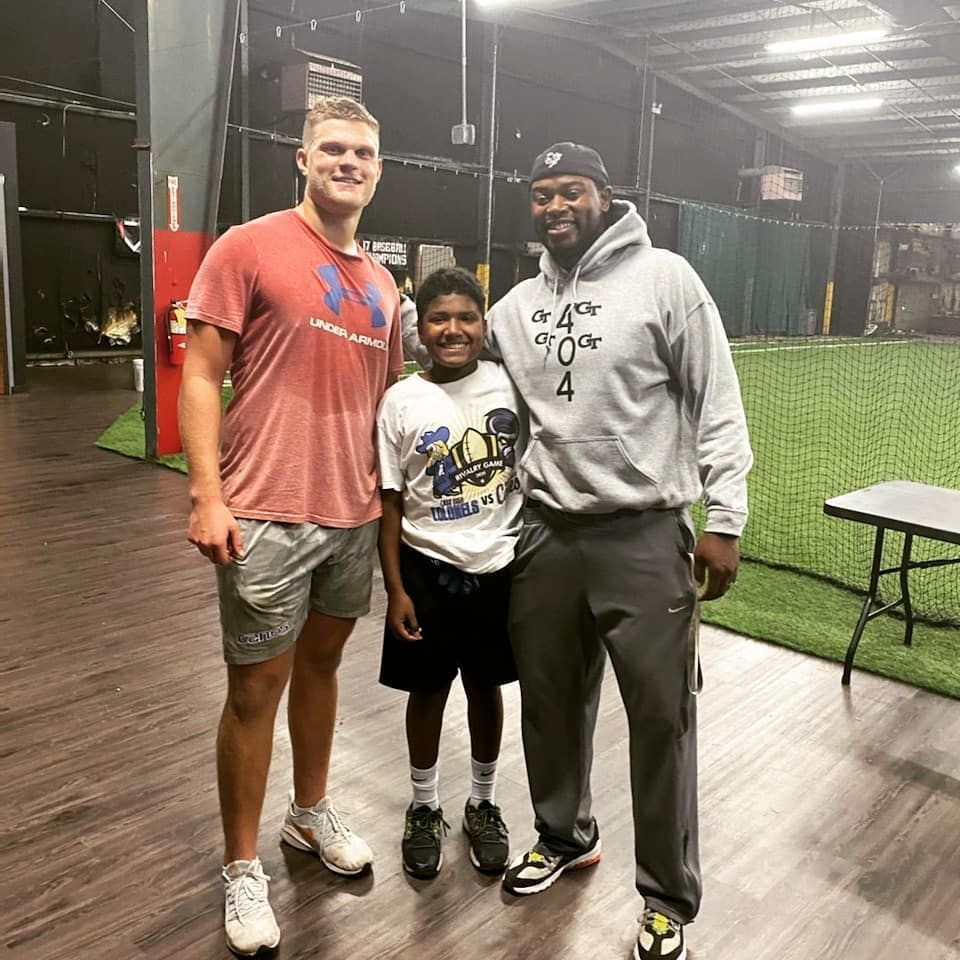 Three people stand side-by-side smiling in an indoor training facility with turf and sports equipment in the background.