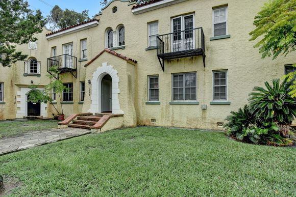 Yellow stucco building with arched entryway, small balconies, and a grassy lawn.