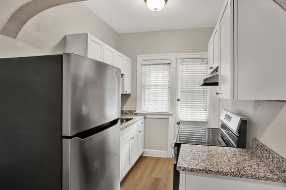 Small kitchen with stainless steel refrigerator, white cabinets, granite countertop, and window with blinds.