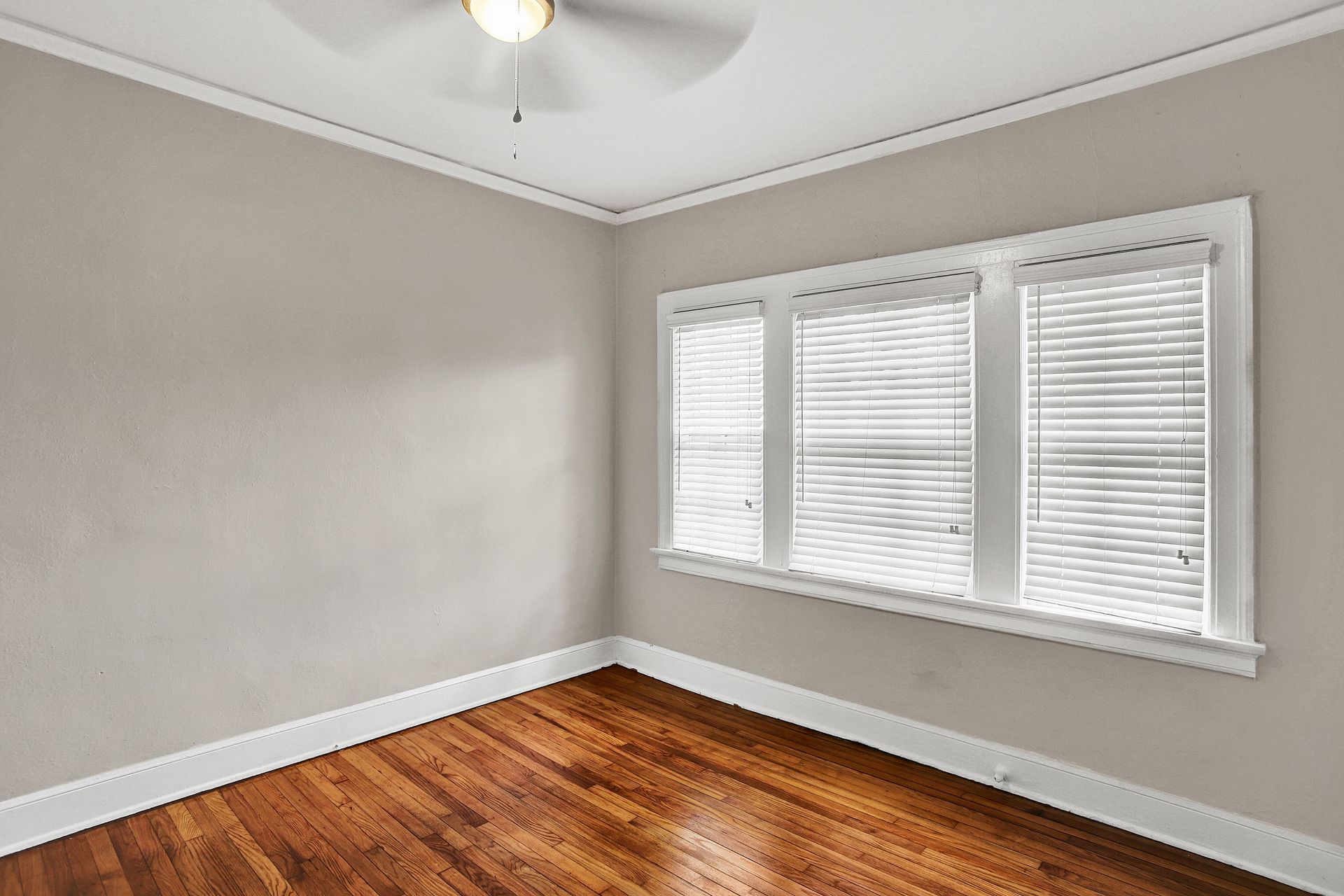 Empty room with hardwood floors, three windows with blinds, and a ceiling fan.