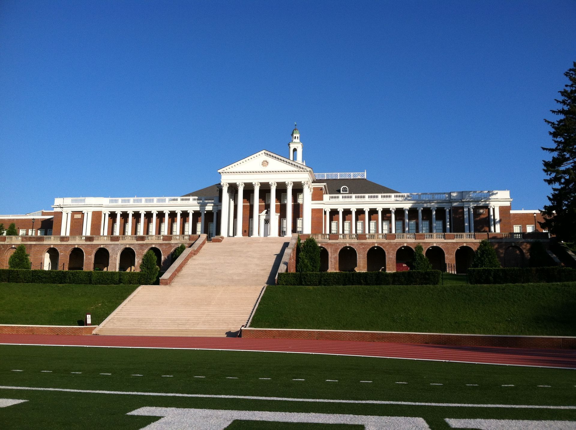 Large brick and white-columned building with a long staircase on a green lawn with a track and football field.