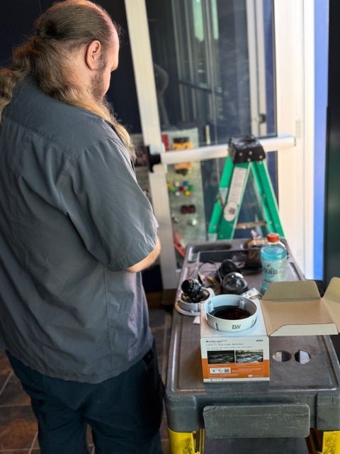 Man in gray shirt works near a ladder. Tools and boxes on a rolling cart. Interior setting, well lit.