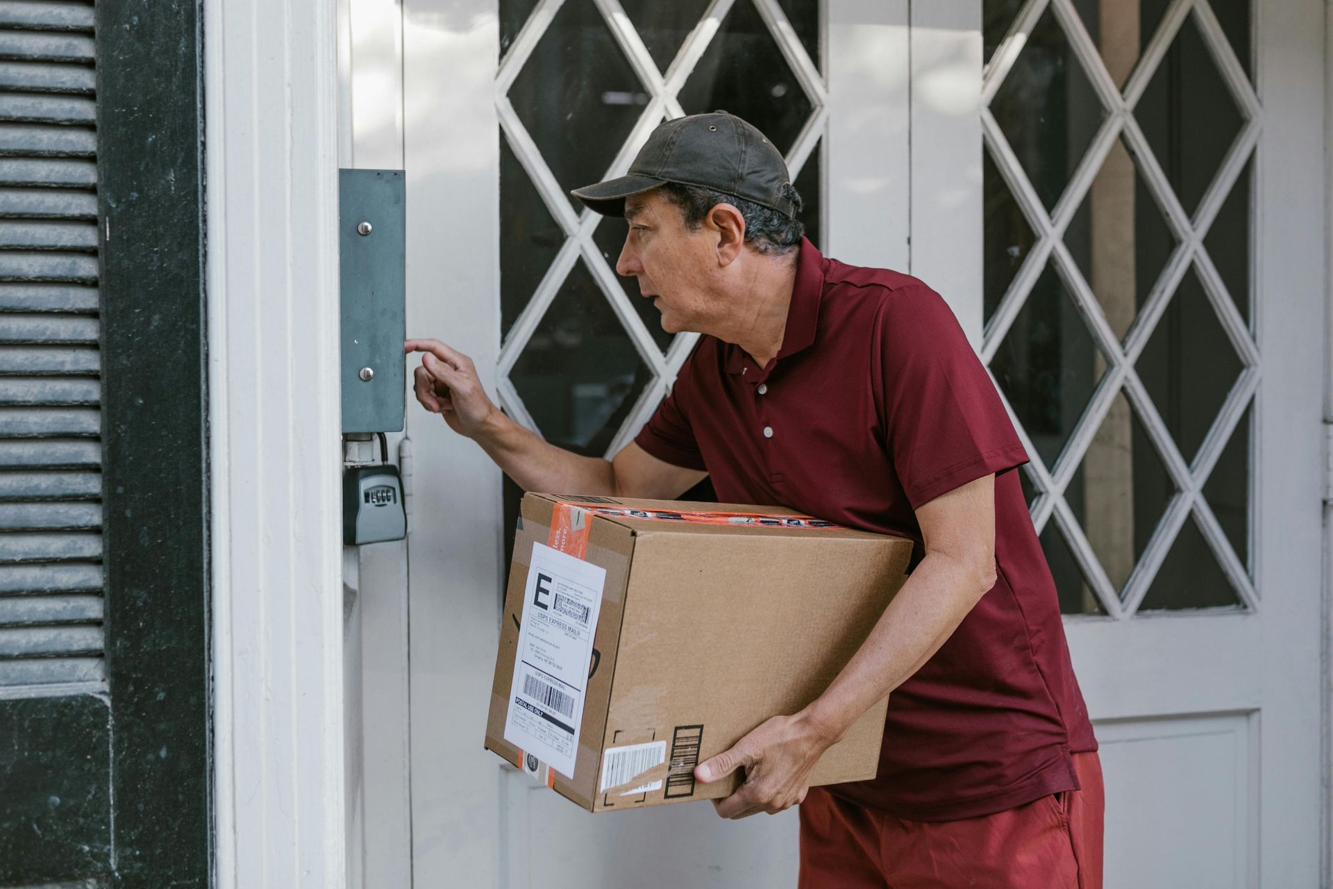 Man in red shirt holding a box, pressing a doorbell on a white door.