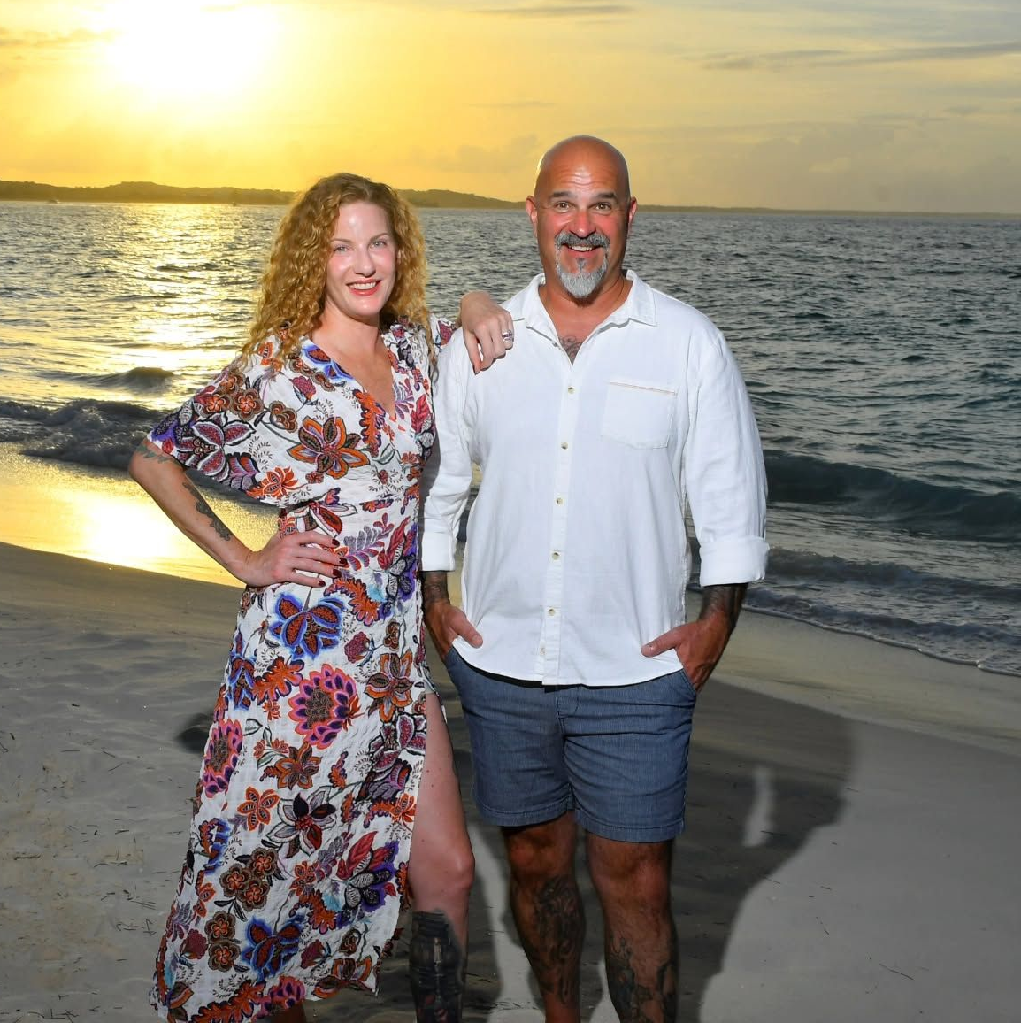 A woman wearing glasses and a gray jacket is smiling on the beach.
