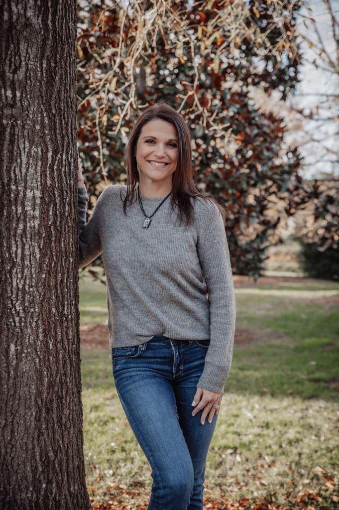 A woman wearing glasses and a gray jacket is smiling on the beach.