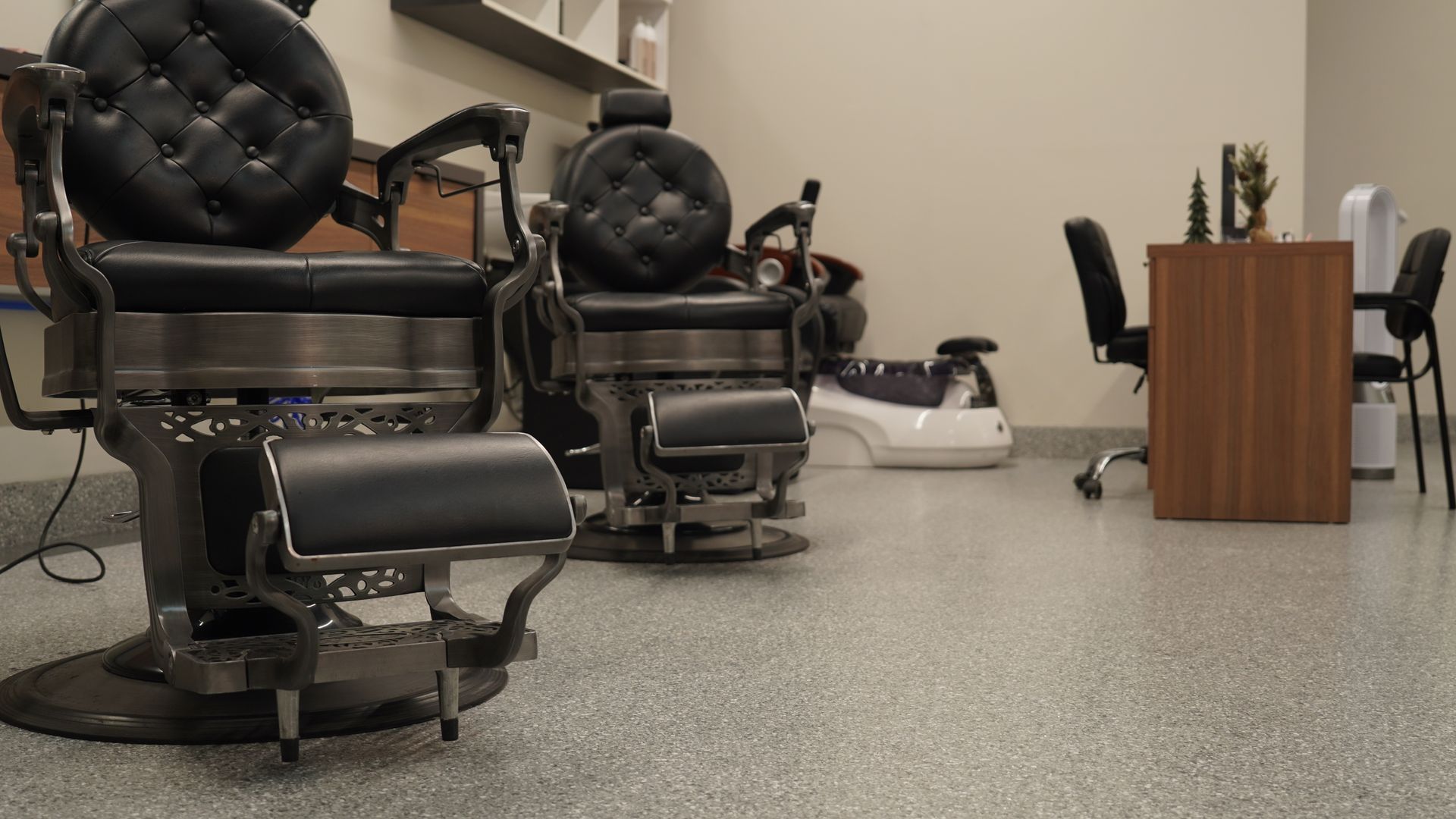 Barber shop interior with two black barber chairs, grey speckled floor, and a small desk.