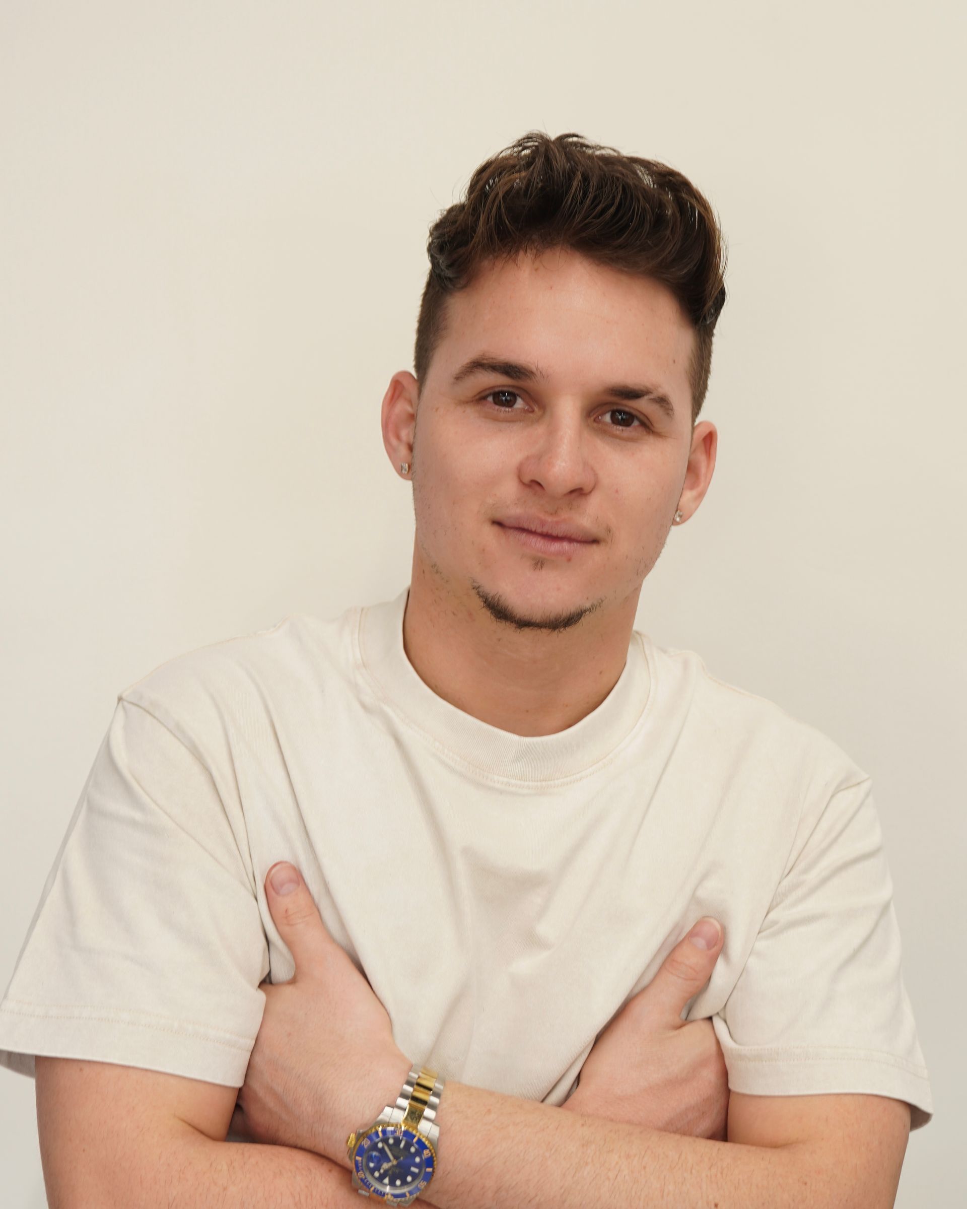 Man with light brown hair, arms crossed, wearing a white t-shirt, and a gold watch.