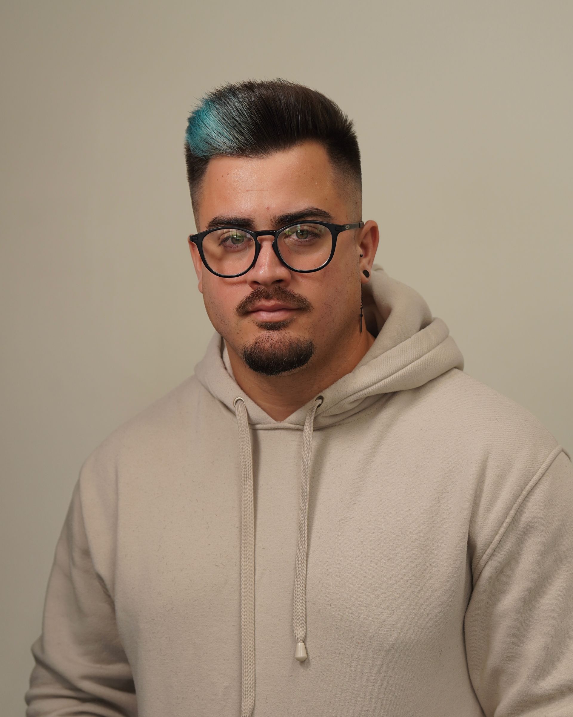 Man wearing glasses and hoodie, with blue-tipped hair, looking forward with a neutral expression.
