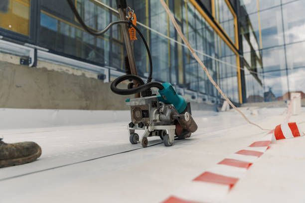Machine welding white roofing membrane on a building's edge. Red/white tape and window reflection visible.
