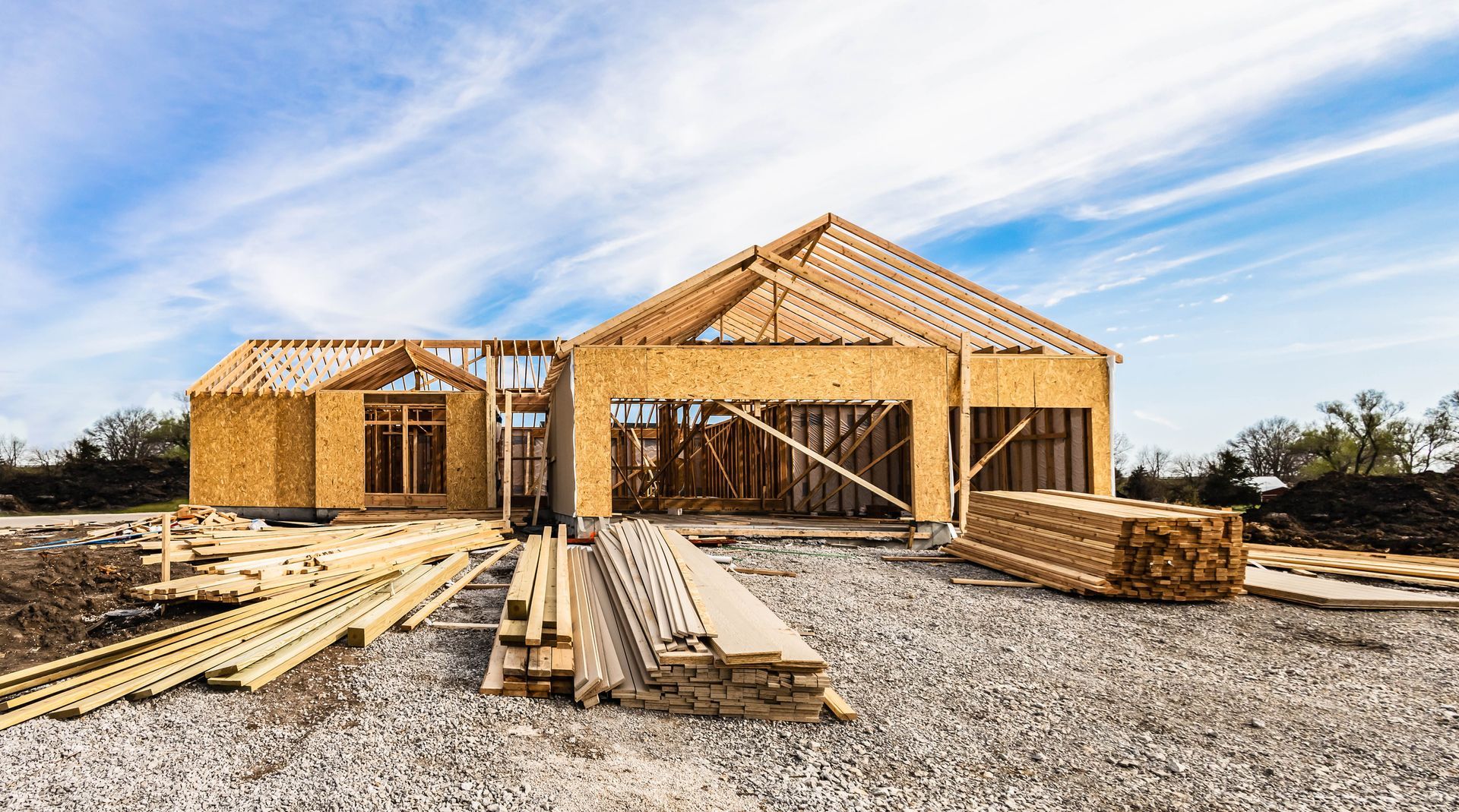 Wood-framed house under construction; beams, lumber and gravel on the ground; blue sky background.