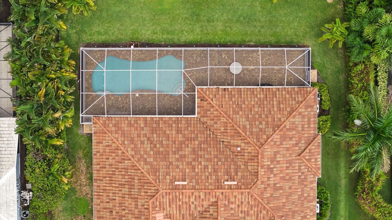 Overhead view: House with a brown roof and a pool with a screen enclosure surrounded by green grass and trees. Overhead view: House with a brown roof and a pool with a screen enclosure surrounded by green grass and trees.