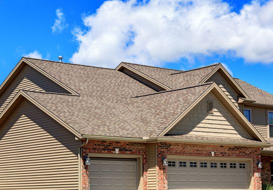 Tan shingled roof on a two-story house with a blue sky and clouds.