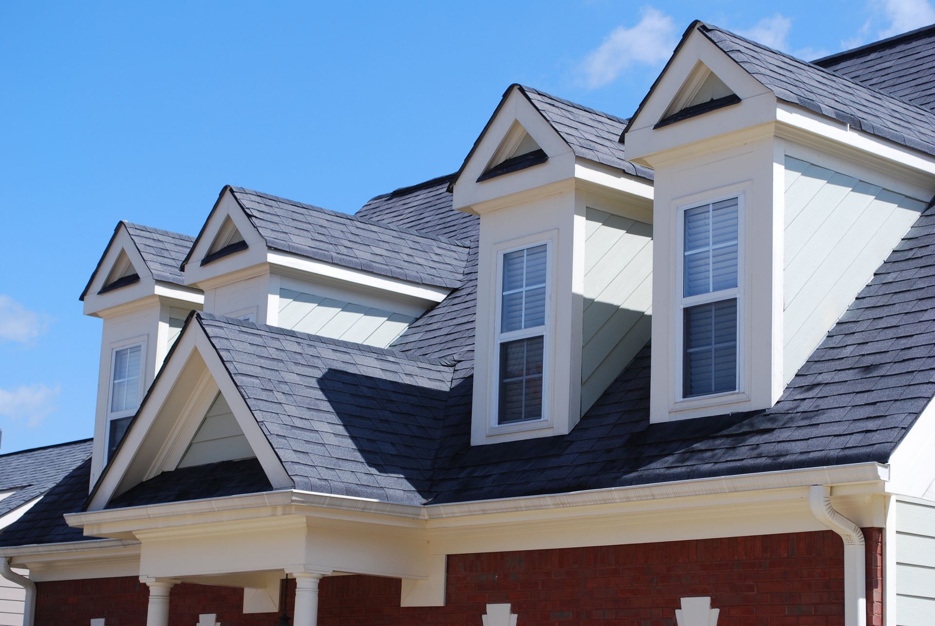 Row of dormers with white trim on a house with a dark roof and red brick facade against a blue sky.