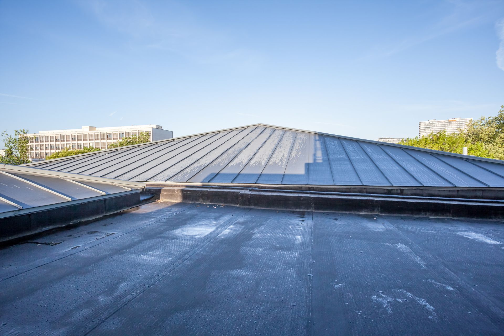 Black flat roof transitions to a sloped, metallic roof, with buildings and a clear sky in the background.