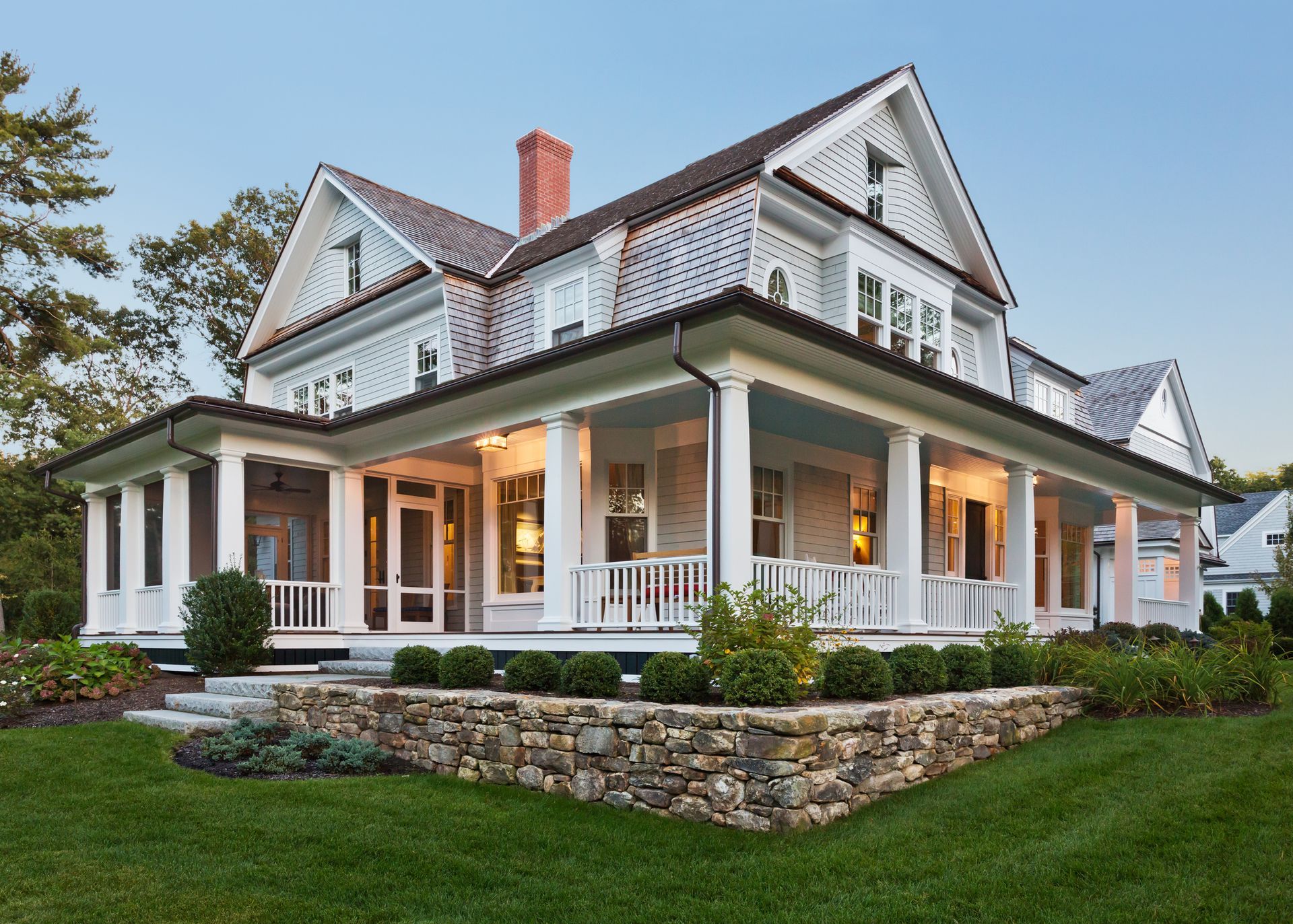 Elegant two-story house with wraparound porch, stone retaining wall, and manicured lawn.