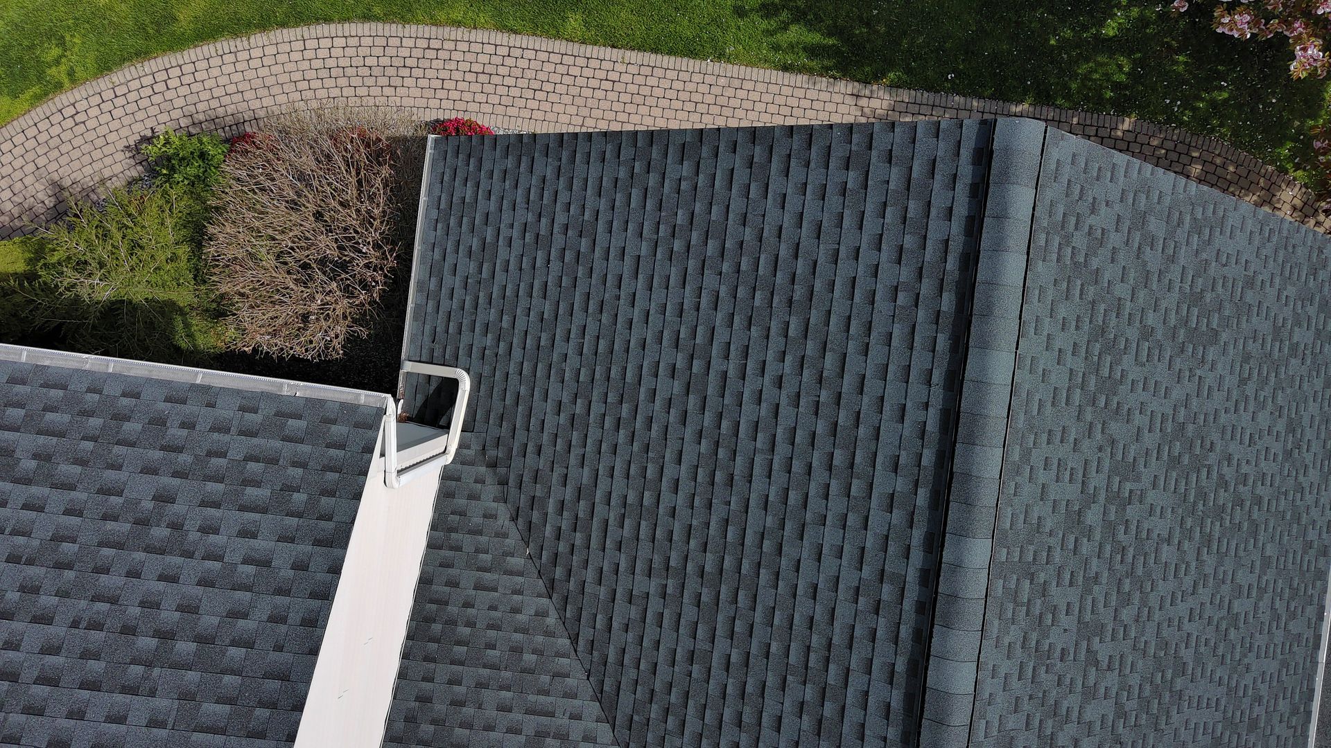 Overhead view of a dark gray shingled roof next to a white chimney and a curved brick pathway.
