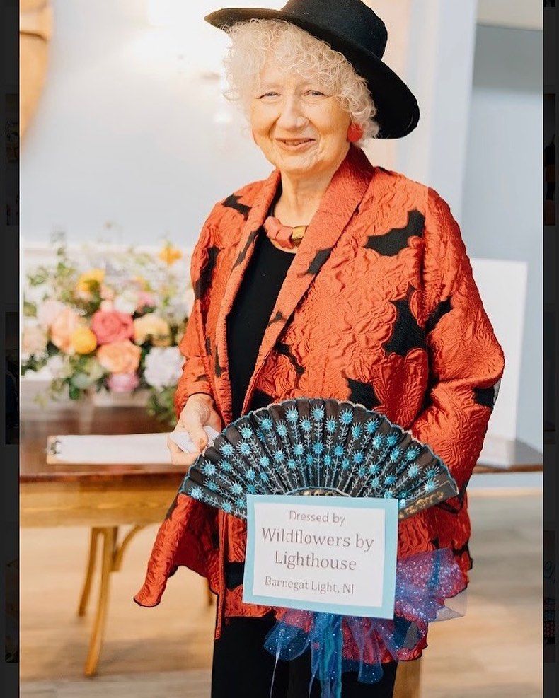 Woman in orange jacket, black hat, holding a fan with a sign, standing near flowers.