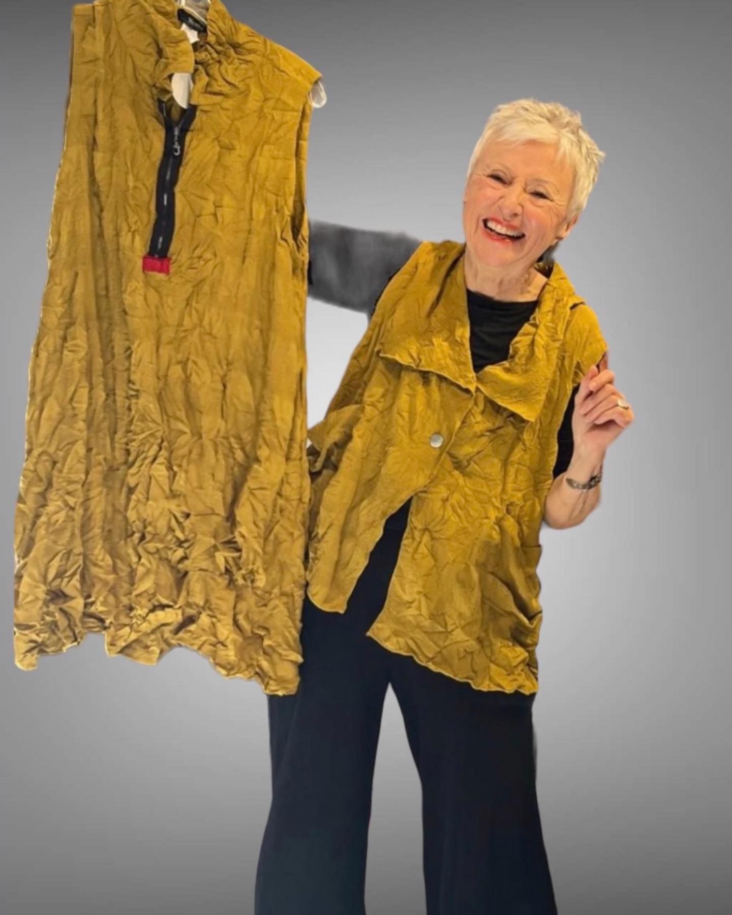 Woman holding up two mustard-colored clothing items, smiling. Gray backdrop.