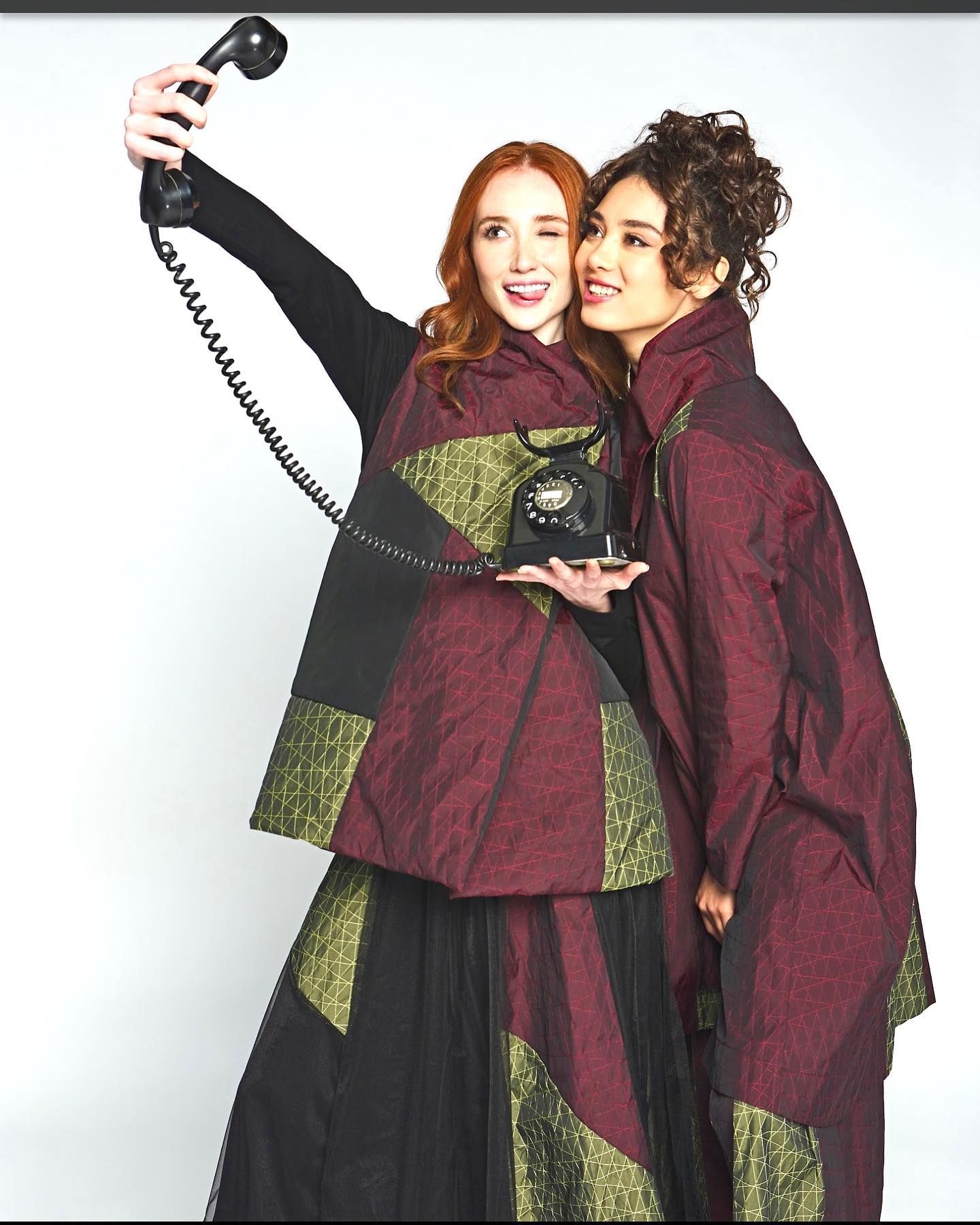Two women pose, one taking a selfie with a vintage phone. They wear red and gold patterned jackets against a white background