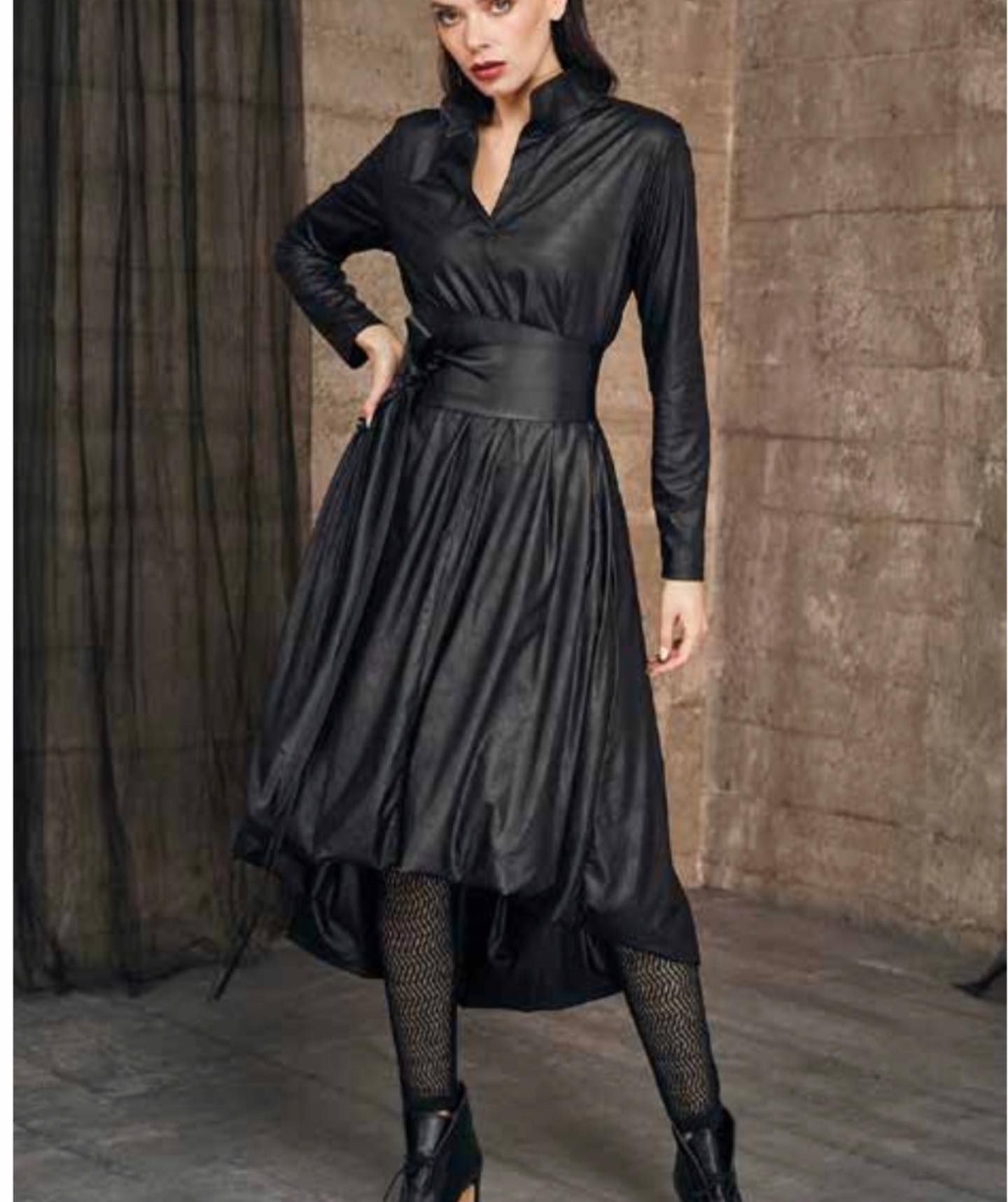 Woman in a black dress with high-low hemline, waist tie, and tights posing indoors.