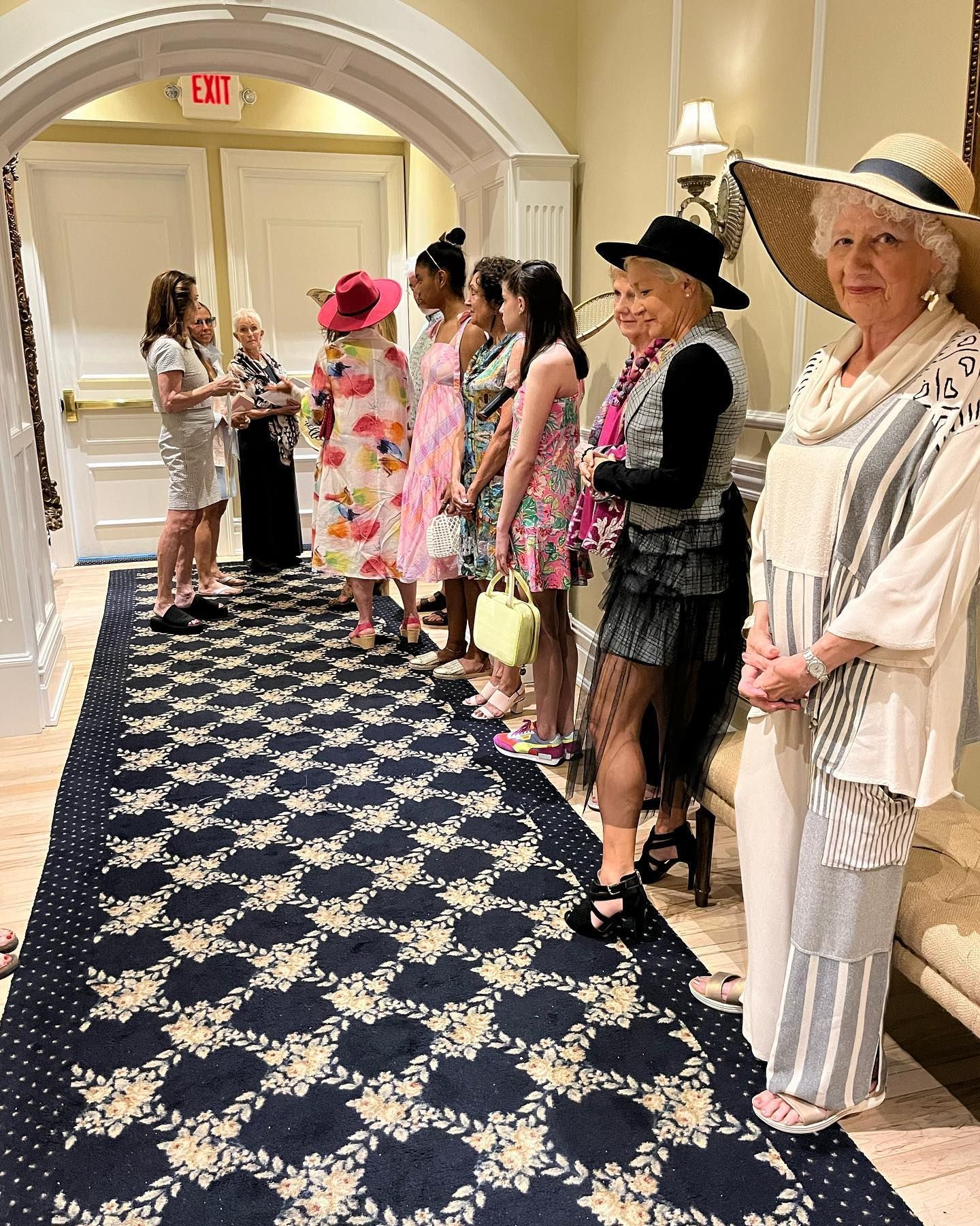 People in stylish hats and dresses line a hallway with a floral rug, likely at an event.