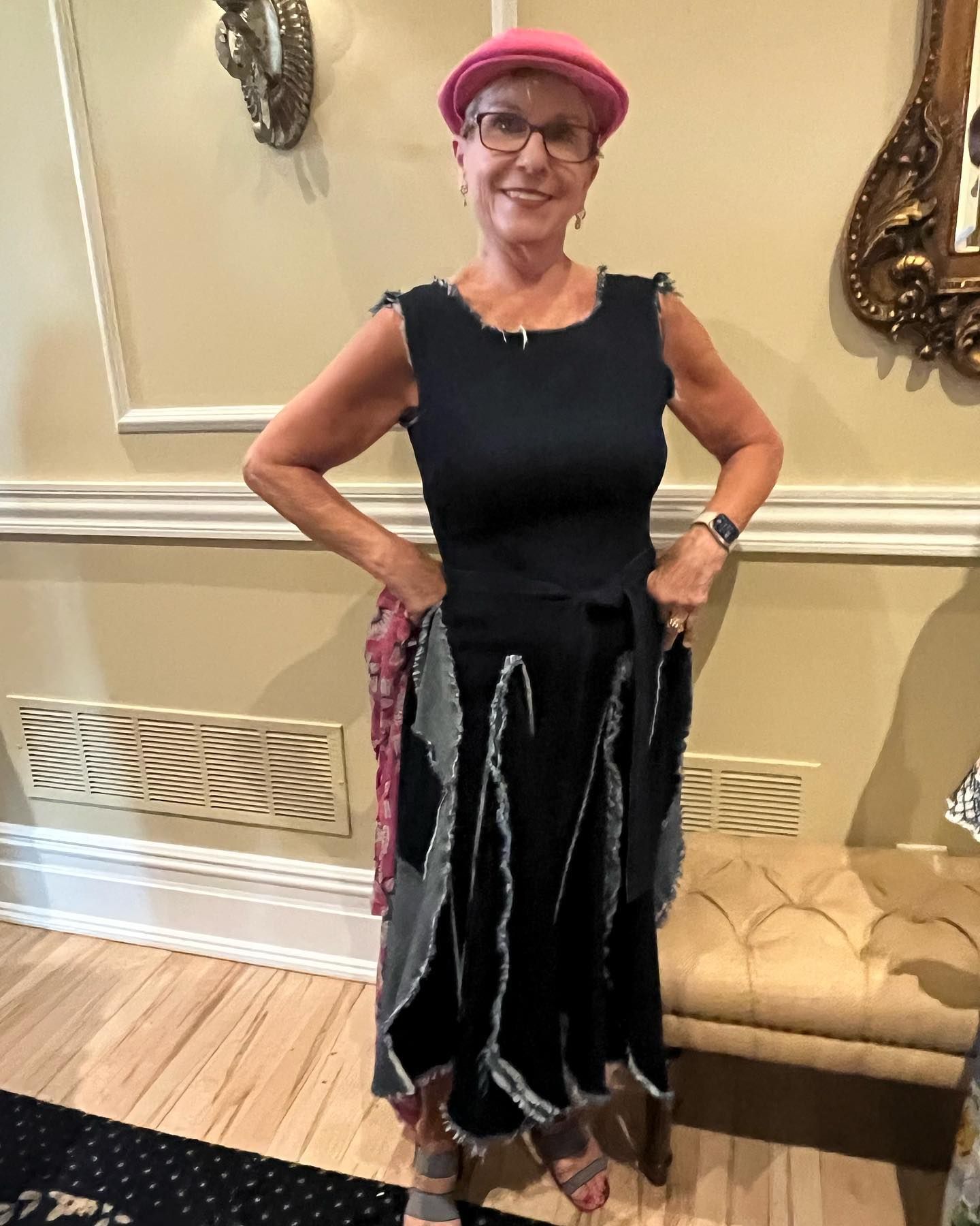 Woman in black dress and pink hat, smiling, hands on hips. Indoors, near a bench and vent.