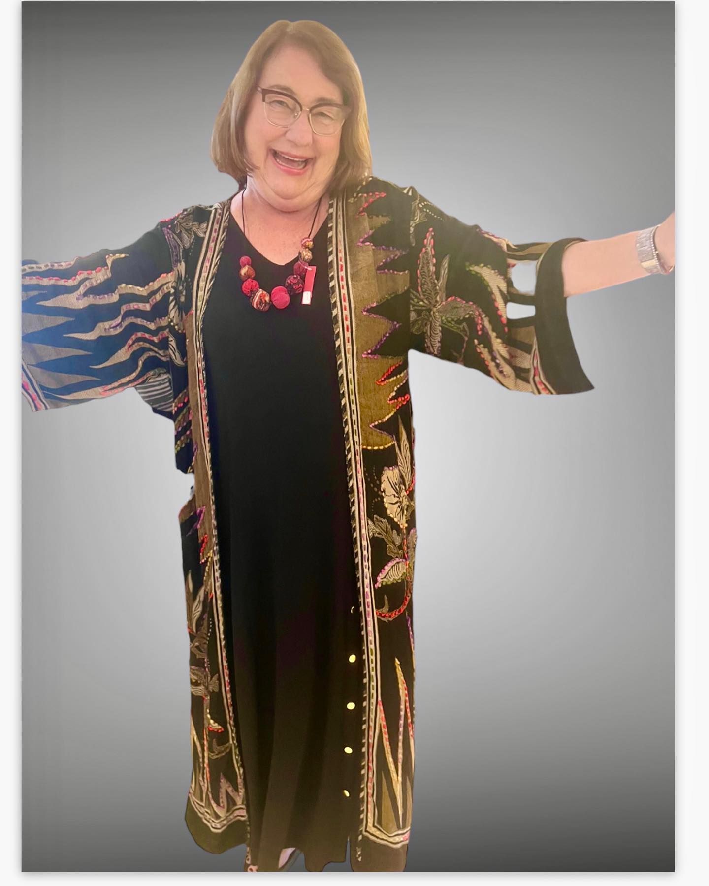 Woman in glasses, black dress, patterned kimono, arms outstretched, smiling.