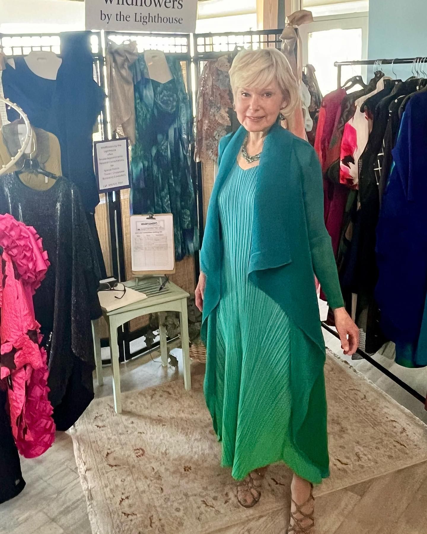 Woman in a teal dress and cardigan stands in a clothing shop. Dresses are on racks.