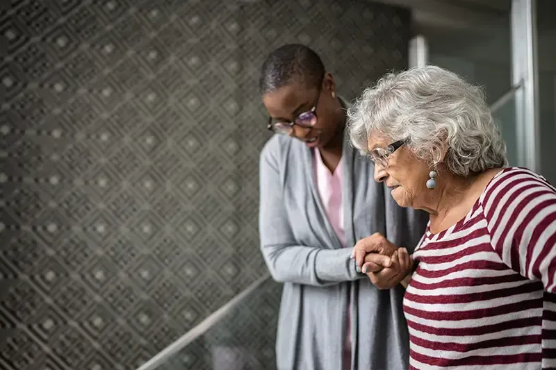 A woman experiencing some signs of dementia is supported by her caregiver as she walks downstairs.