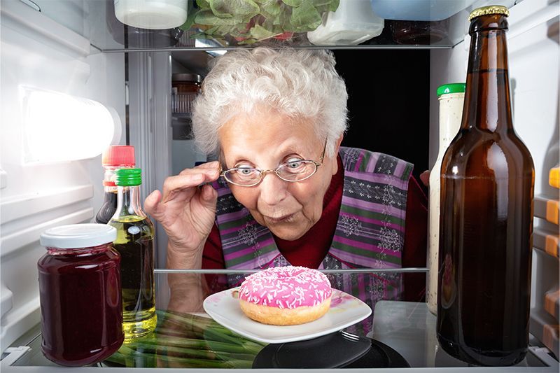 An older woman experiencing food addiction in older adults peers into the fridge at a frosted donut.