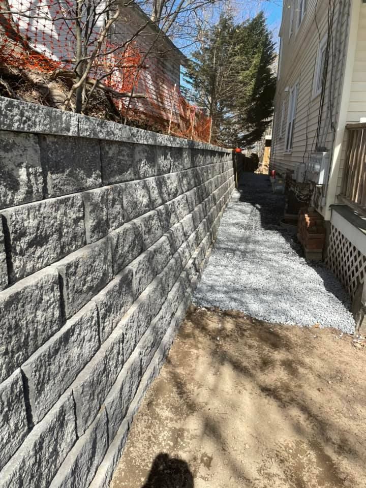 Gray retaining wall next to a narrow gravel path and building. Soil in the foreground.