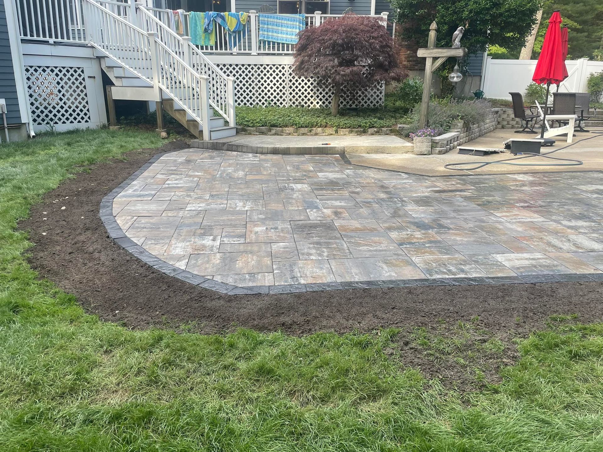 Paver patio with a dark border, bordered by fresh soil and grass, near a deck.