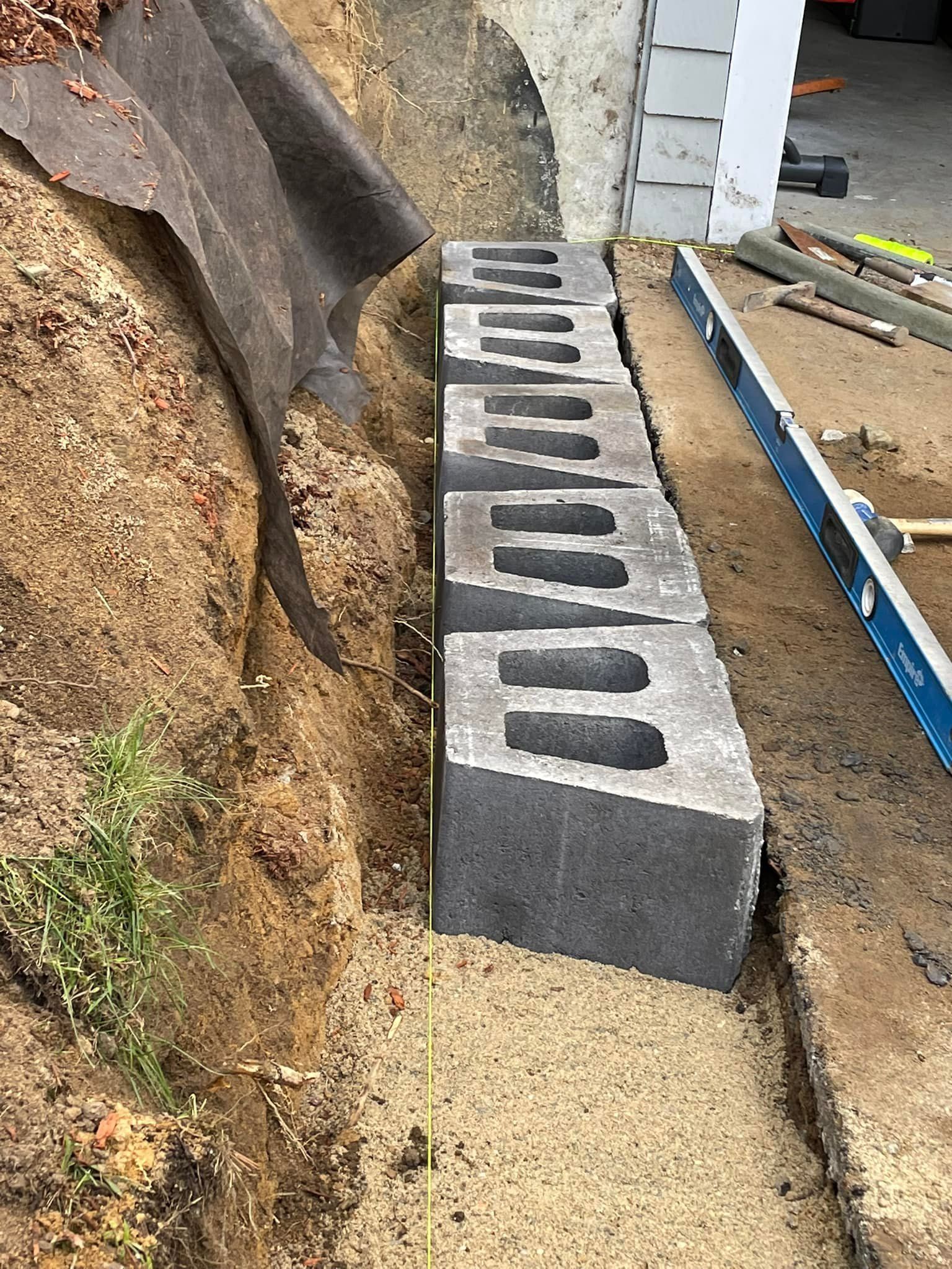Cinder block retaining wall under construction next to a building. Soil and weed barrier visible.