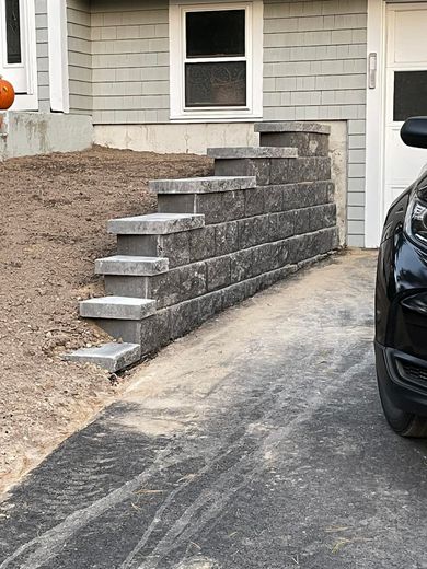 Gray retaining wall with steps next to a driveway and building. Unpaved dirt area is to the left.