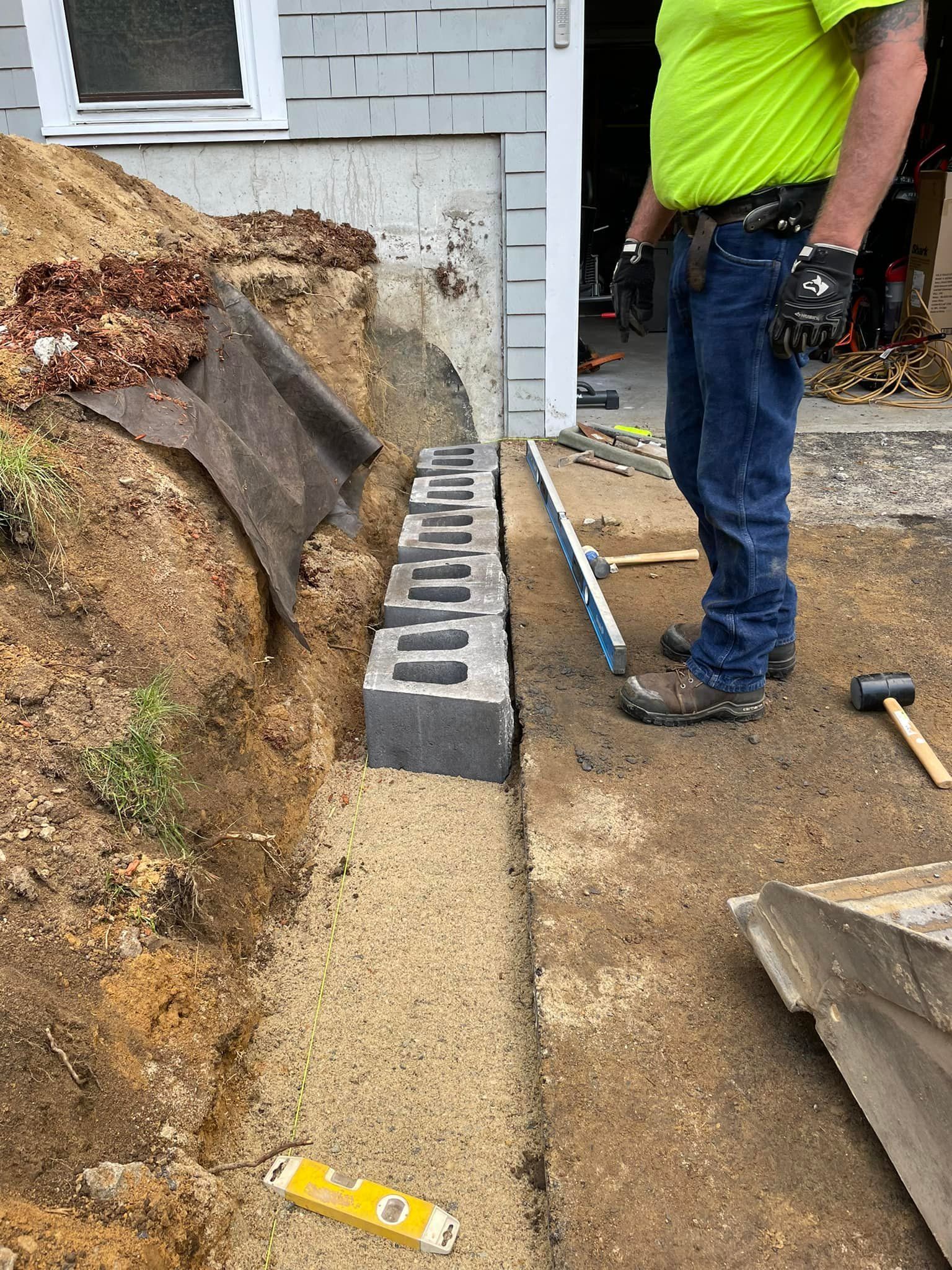 Construction worker building a retaining wall with concrete blocks near a building's foundation.
