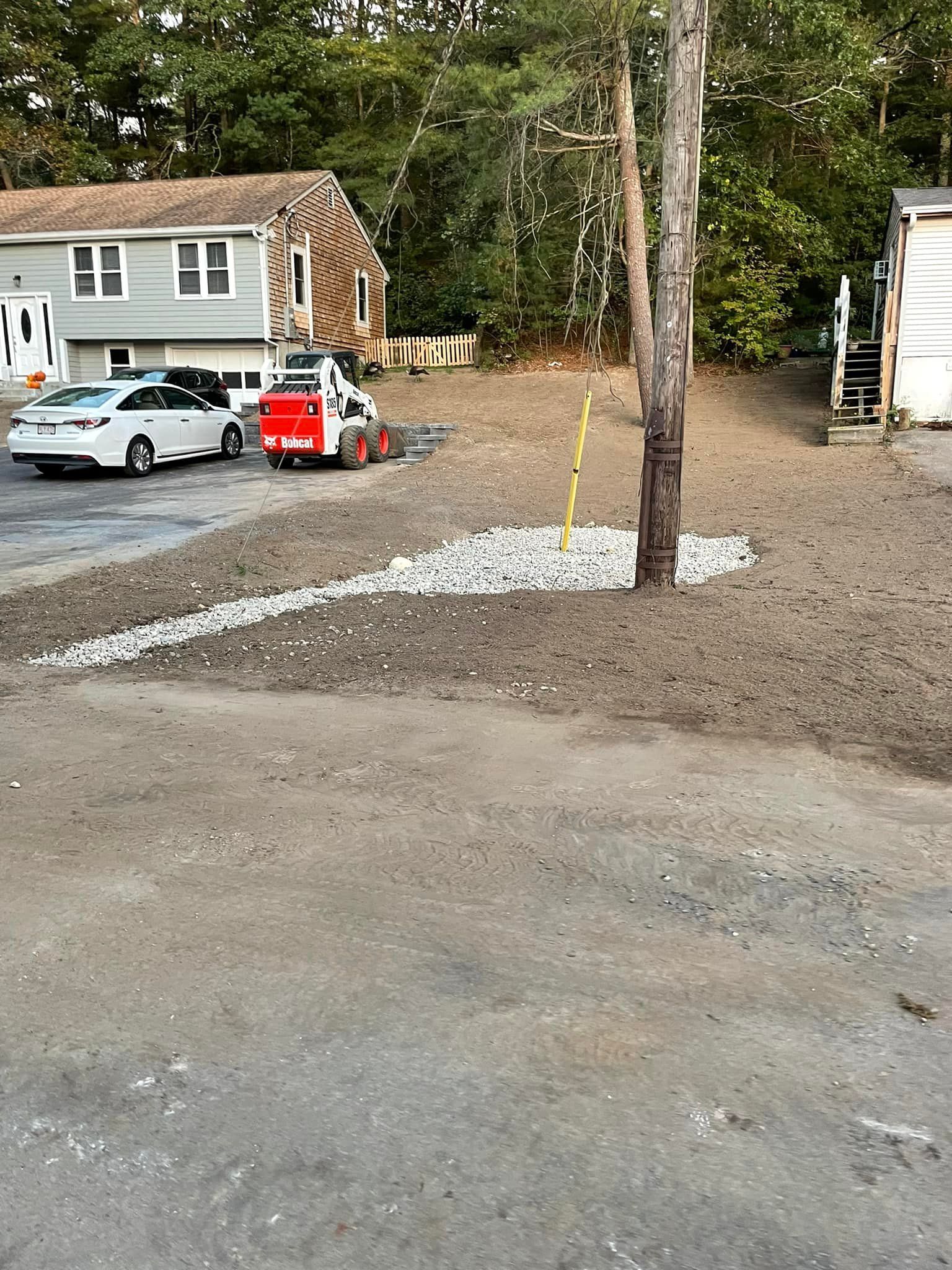 A construction site with a small excavator near a tree, rocks, and a building.