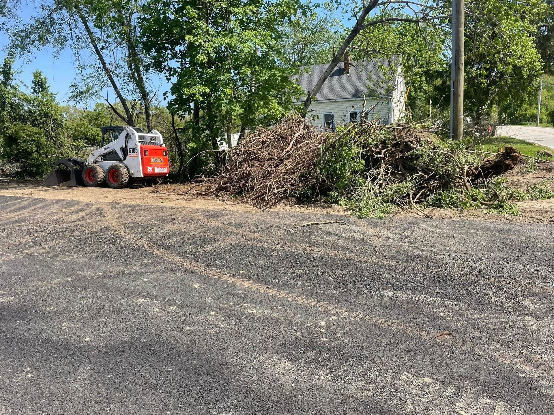 Bobcat clearing brush next to a white house with a dark roof on a sunny day.