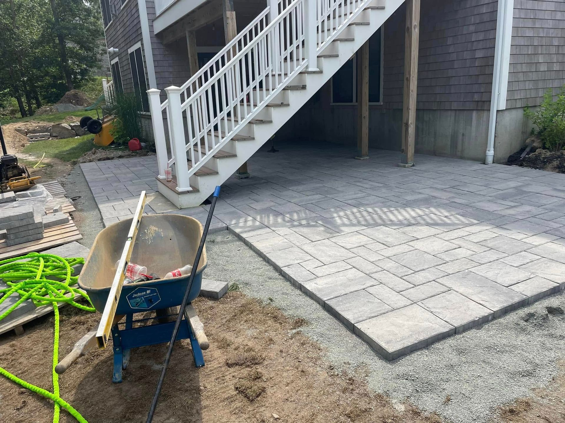 Patio pavers being installed under a white staircase next to a house; wheelbarrow and hose in foreground.