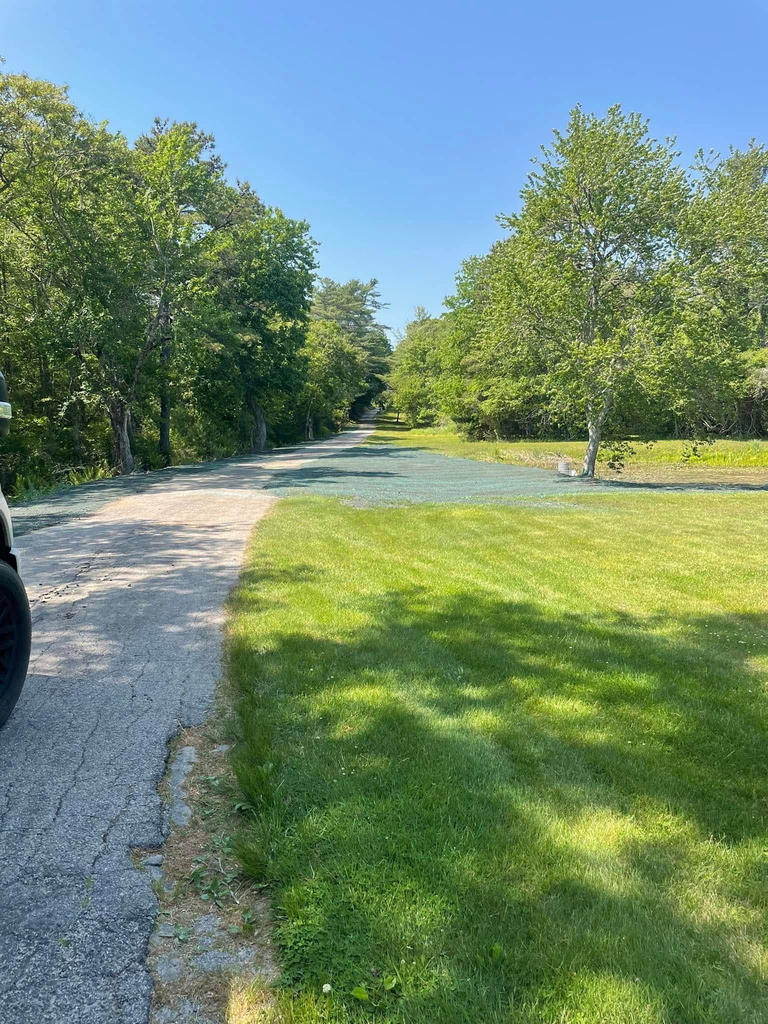 Gravel road alongside green grass and trees under a clear blue sky.