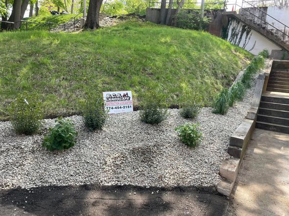 Landscaped yard with small shrubs, gravel, and grassy hill, a sign posted.