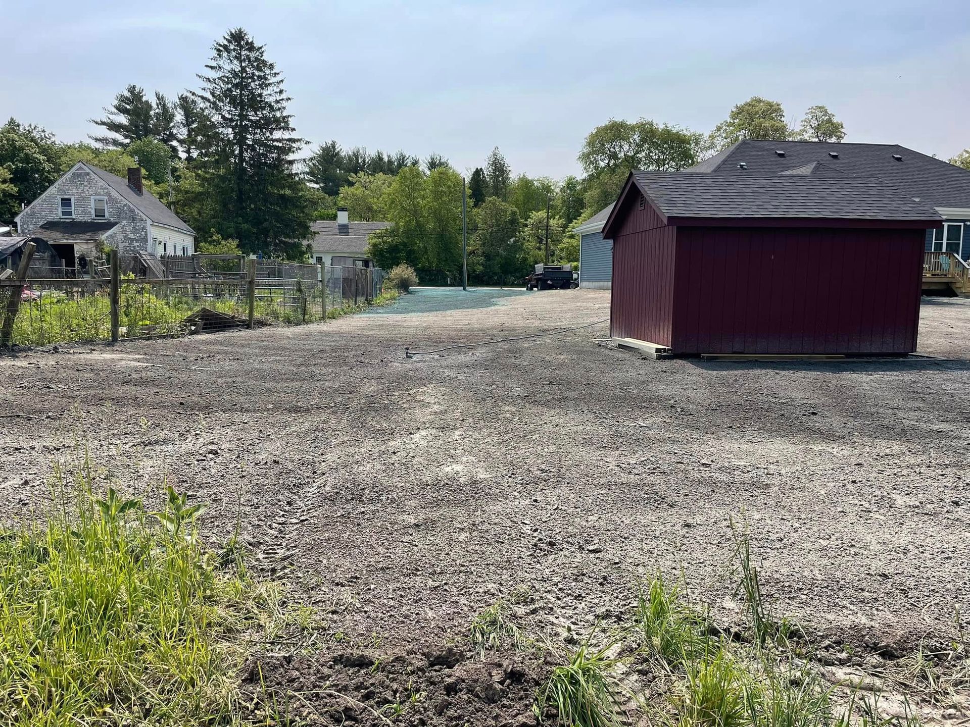 Gravel-covered lot with red shed, fence, and houses in background under a sunny sky.