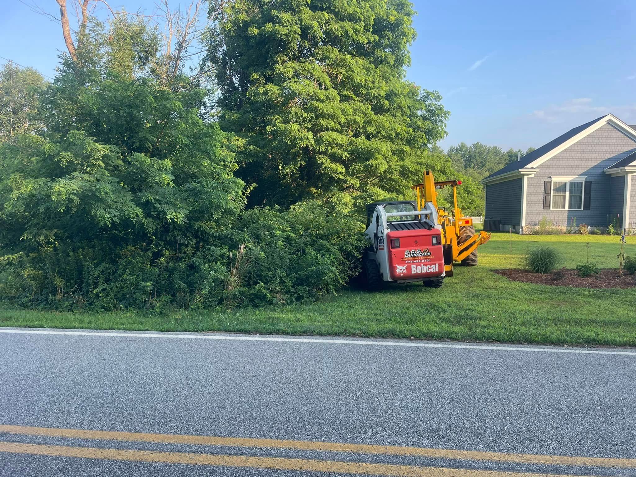 Red and yellow construction equipment trimming bushes near a gray house next to a road with double yellow lines.