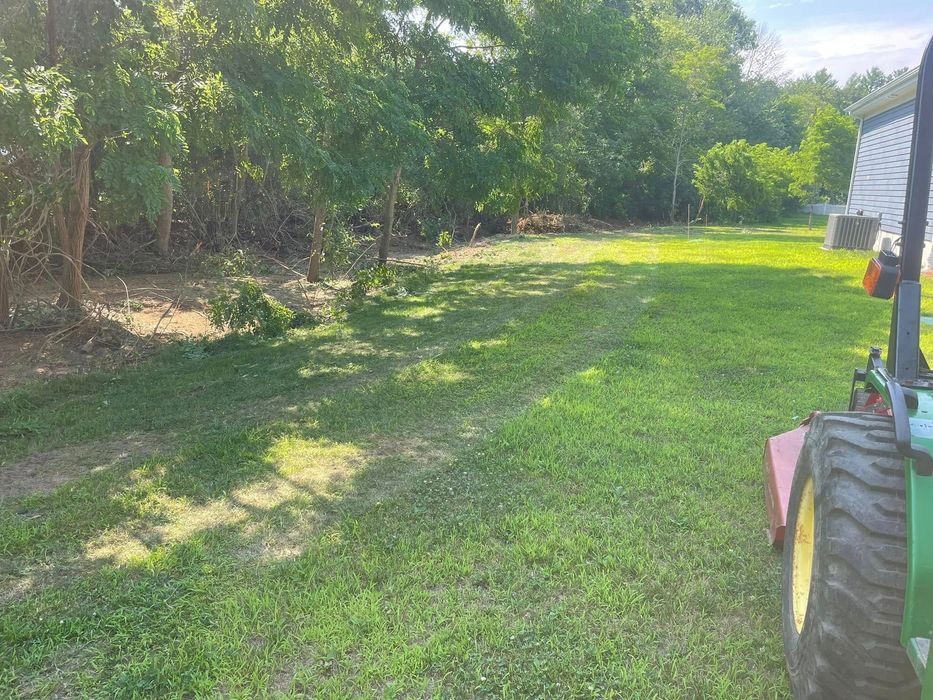 Lawn being mowed with a tractor. Green grass, trees, and a house in the background. Bright sunlight.