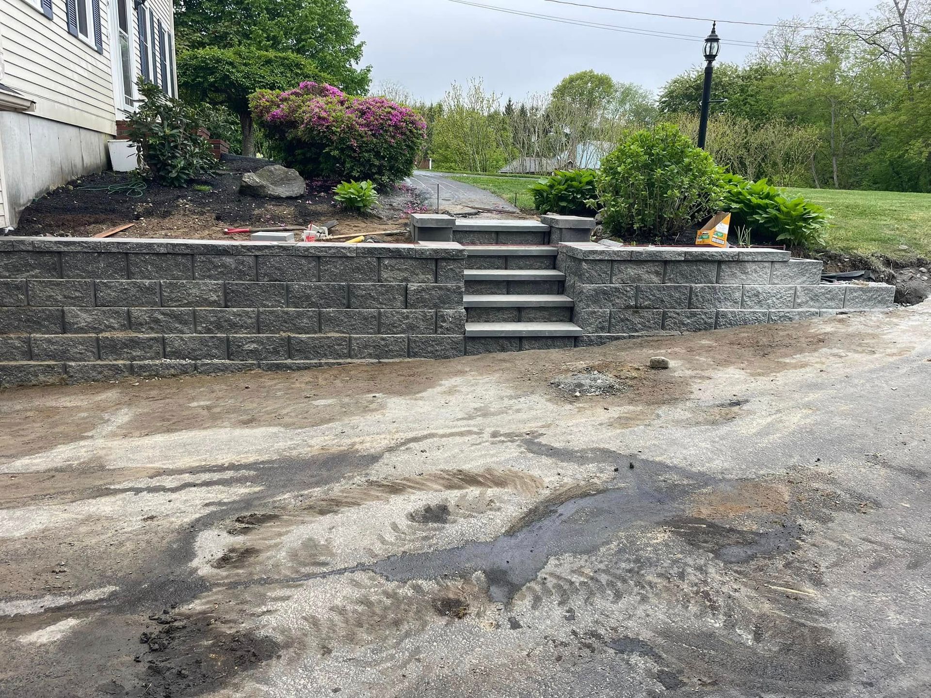 Retaining wall with steps leading to a yard with bushes and flowers. Gray blocks and paving stones.