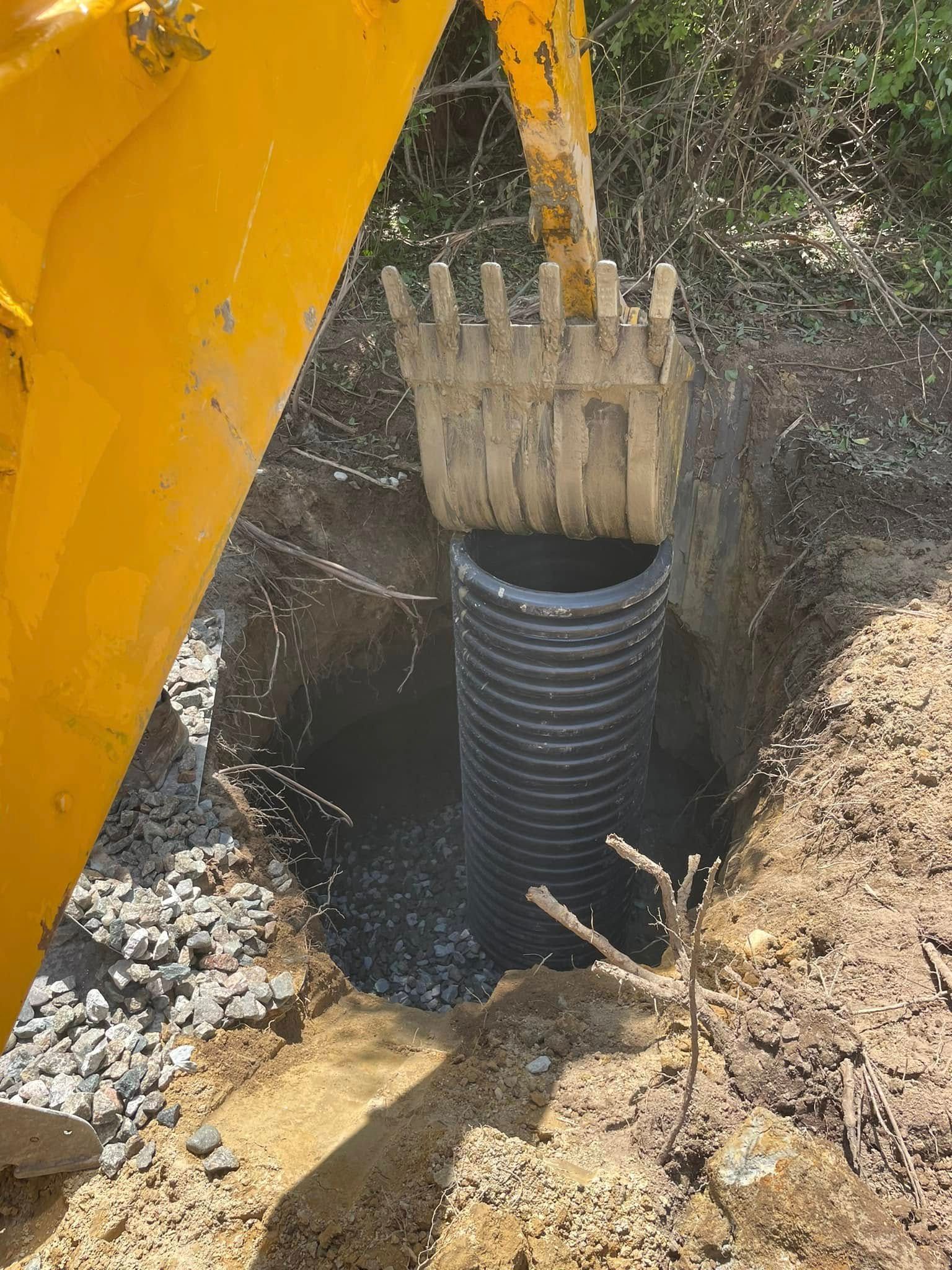 Yellow excavator placing a black corrugated pipe in a dug trench.