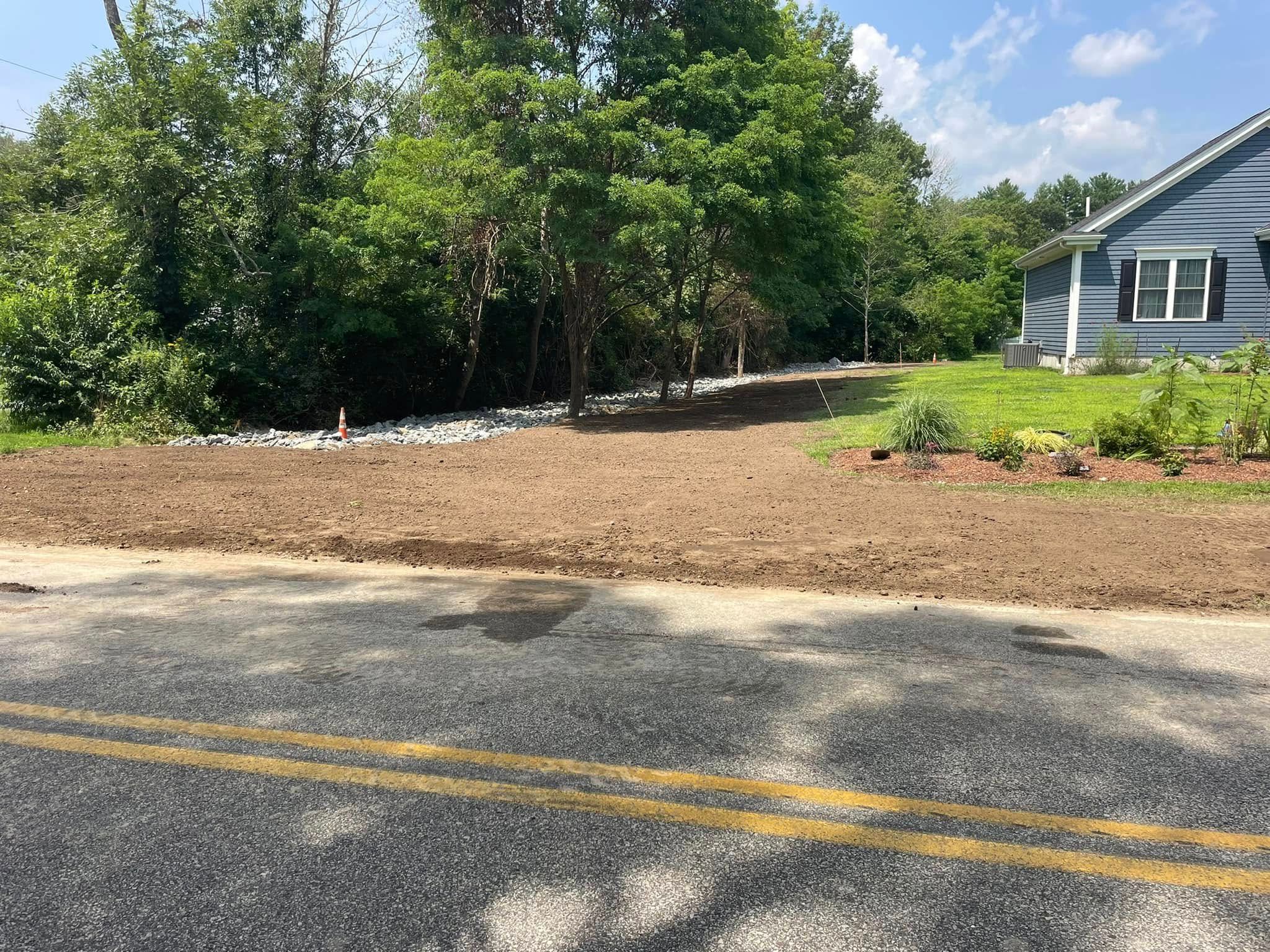 A dirt patch borders a road, leading to a house with trees and a blue exterior on a sunny day.