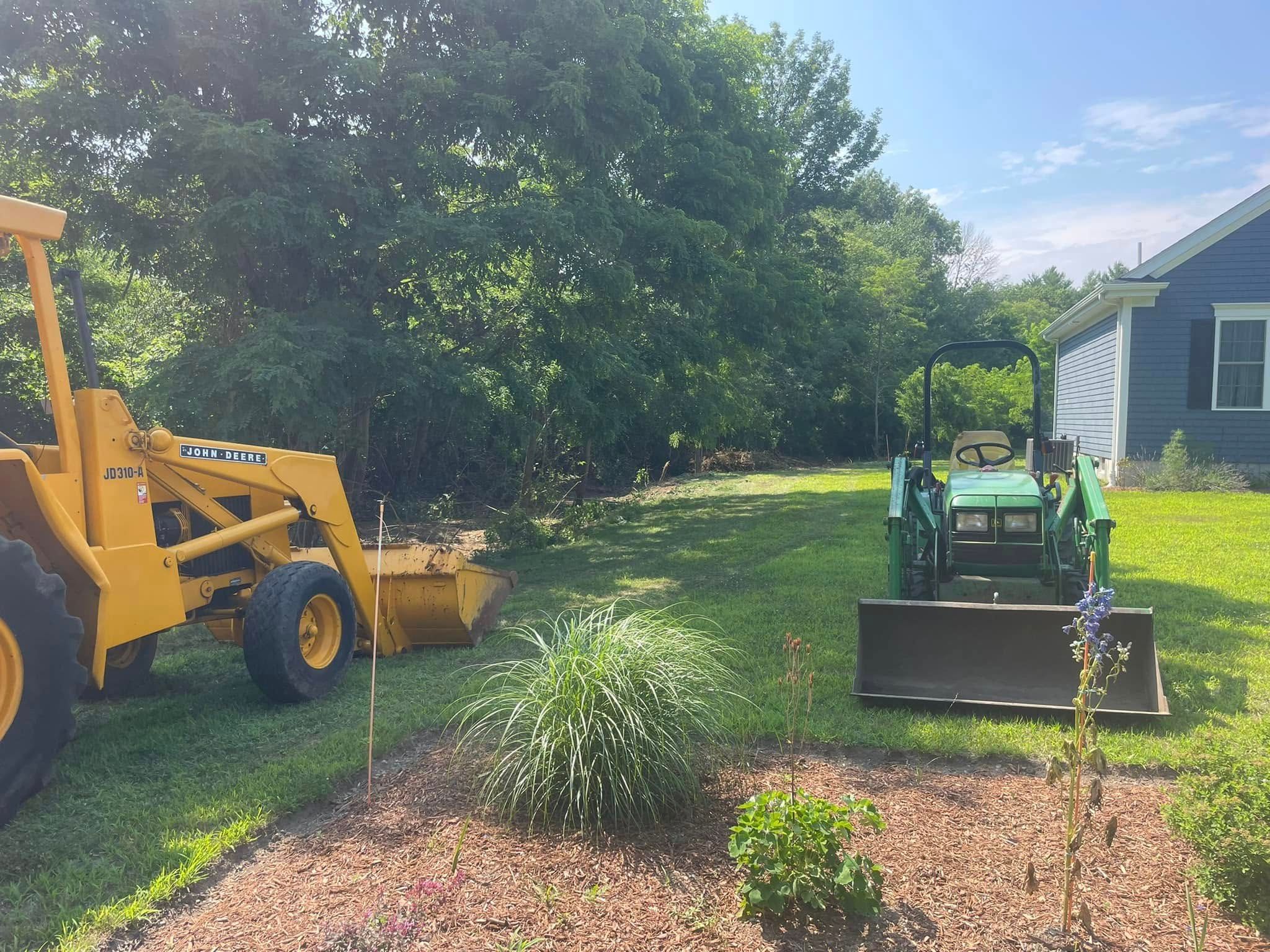 Two tractors on a grassy lawn with a treeline and a blue house. One tractor is yellow, the other green.
