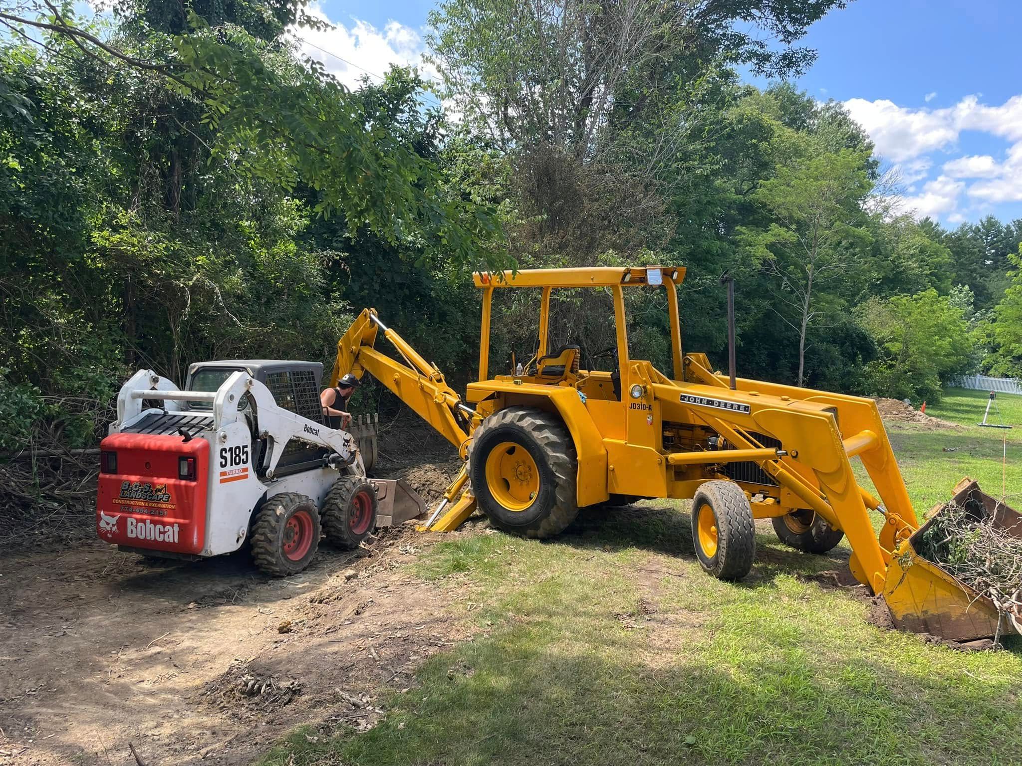 A Bobcat skid steer and yellow backhoe digging near trees in a yard.