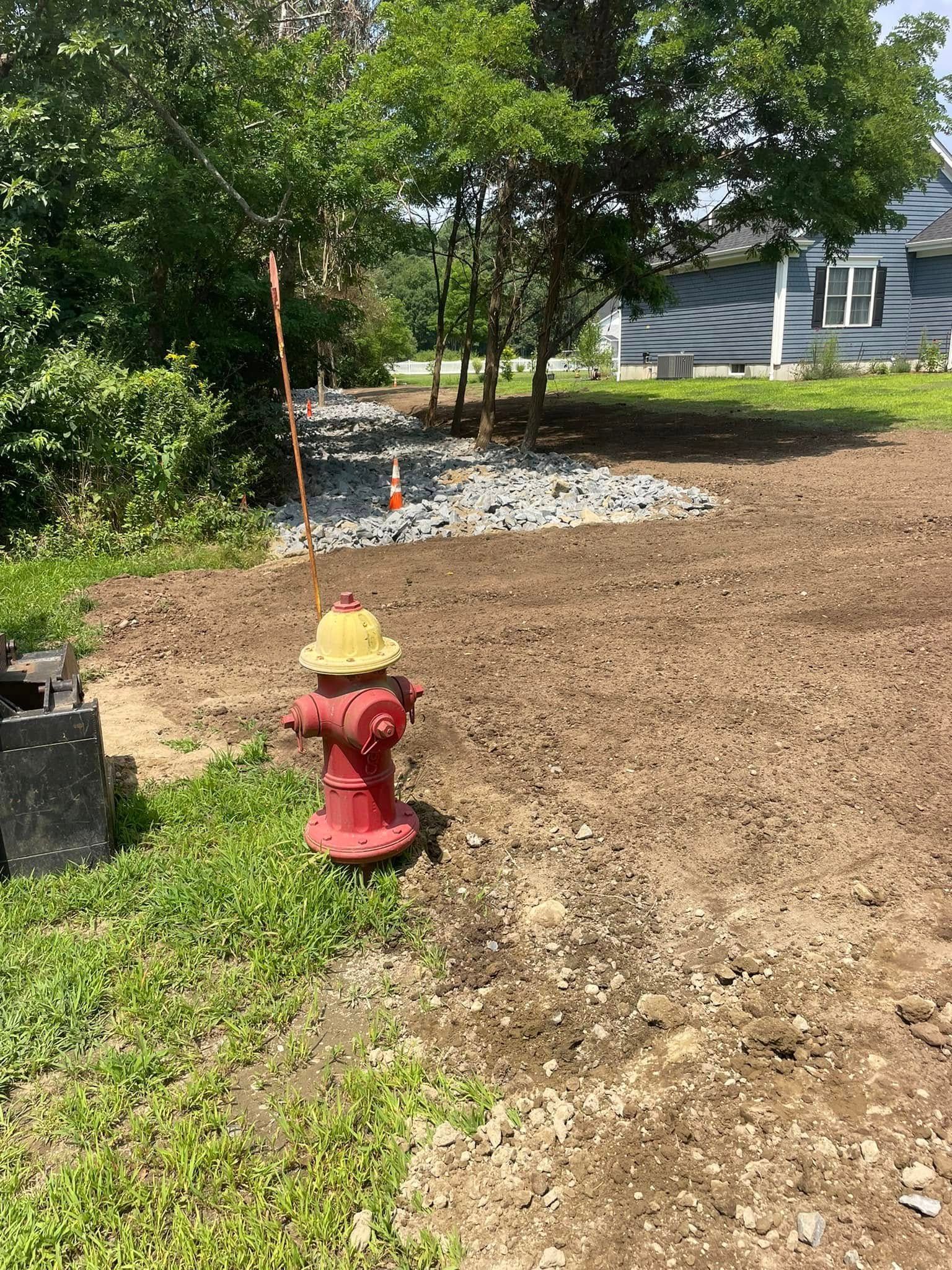 Red fire hydrant in a grassy area with dirt and a rocky patch; a house is in the background.