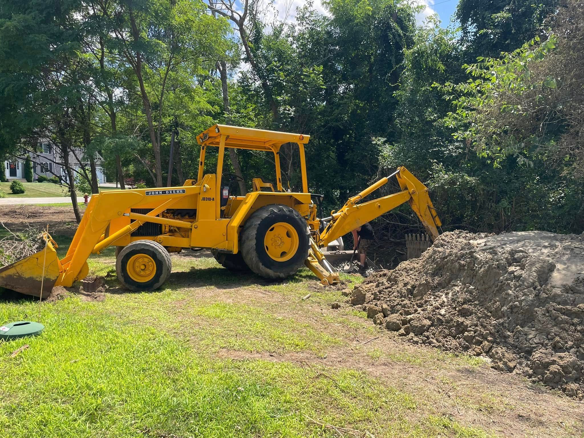 Yellow tractor with a backhoe digging into a pile of dirt in a grassy area, trees in the background.